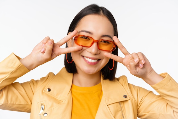 Close up portrait of young asian woman stylish girl in sunglasses showing peace vsign and smiling standing over white studio background