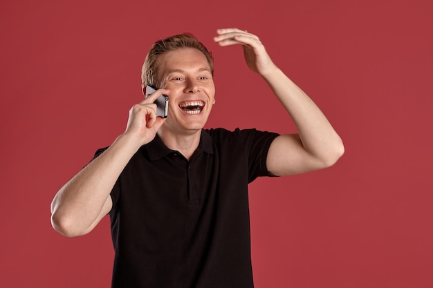 Крупным планом портрет молодого спортивного рыжего мужчины в стильной черной футболке смеется и разговаривает по смартфону, позируя на розовом студийном фоне. Выражение лица человека. Искренние эмоции