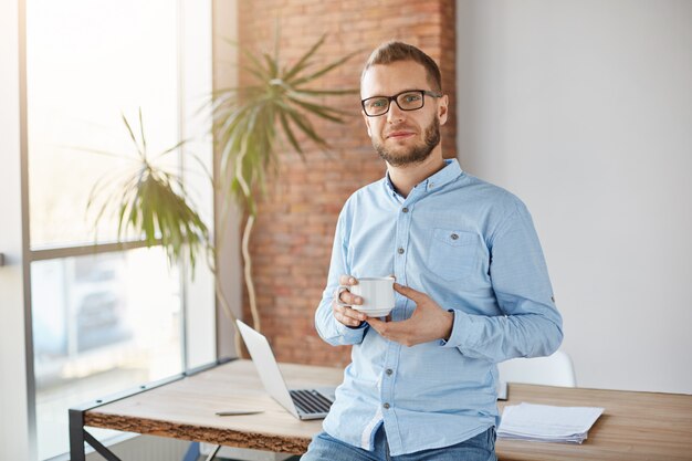 Close up portrait of young attractive company founder in glasses and casual outfit, standing in personal office