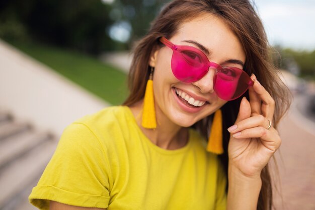 Крупным планом портрет молодой привлекательной улыбающейся женщины, весело проводящей время в городском парке, позитивной, счастливой, в желтом топе, серьгах, розовых солнцезащитных очках, тенденции моды в летнем стиле, стильных аксессуарах, красочных