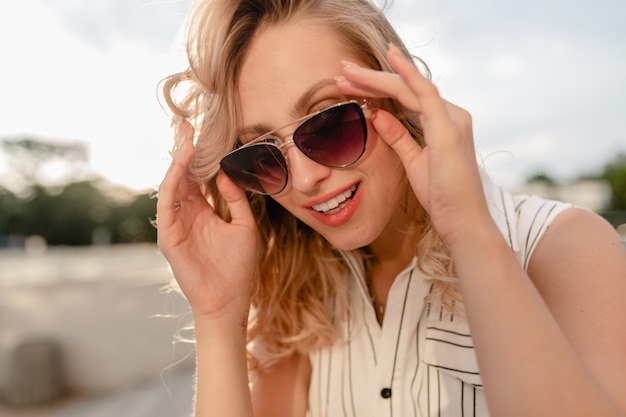 Close-up portrait of young attractive stylish blonde woman in city street in summer fashion style dress holding sunglasses
