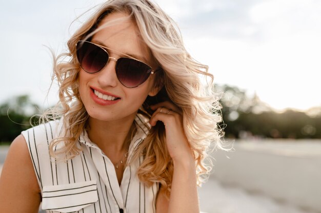 Close-up portrait of young attractive stylish blonde woman in city street in summer fashion style dress wearing sunglasses