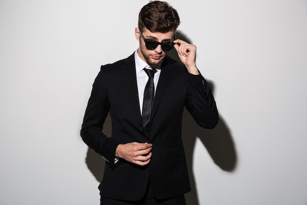 Close-up portrait of young bearded man in black suit touching his sunglasse