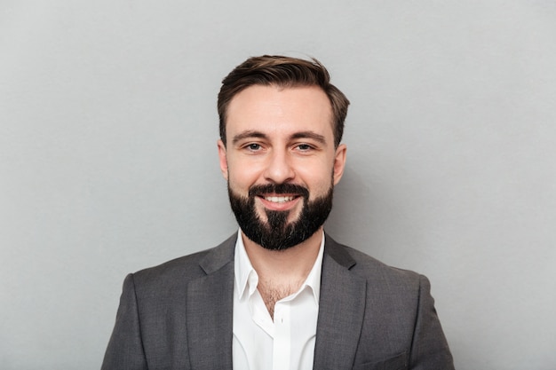 Close up portrait of young bearded man in white shirt and jacket posing on camera with broad smile, isolated over gray