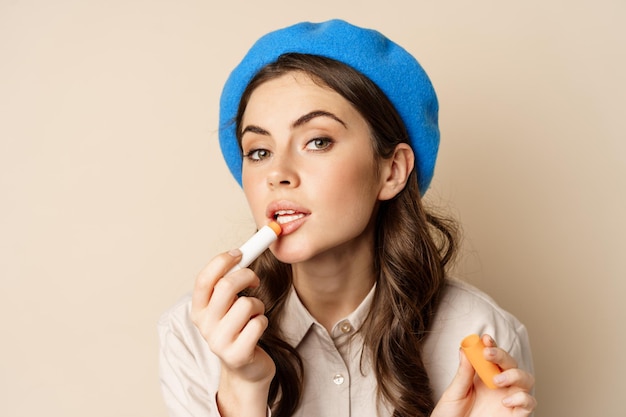 Free photo close up portrait of young beatufiul woman looking in mirror and fixing her makeup, put on lipstick, standing over beige background.