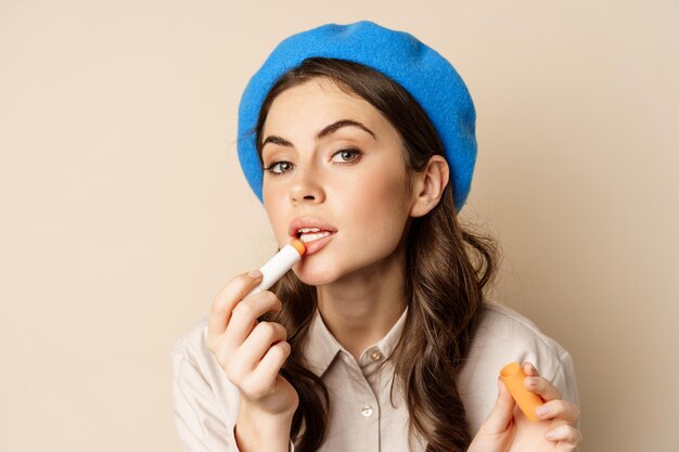 Close up portrait of young beatufiul woman looking in mirror and fixing her makeup, put on lipstick, standing over beige background.