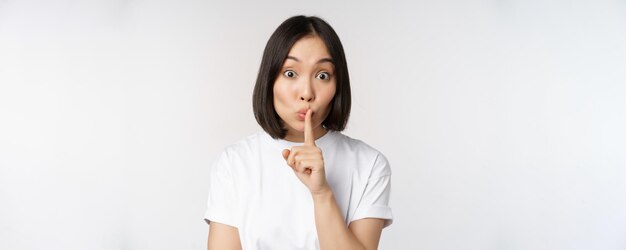 Close up portrait of young beautiful asian girl shushing has secret keep quiet silence gesture press finger to lips standing in tshirt over white background