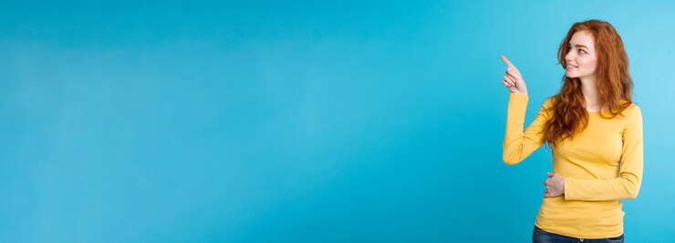 मुफ़्त फोटो close up portrait young beautiful attractive redhair girl happy with something and pointing finger blue pastel background copy space