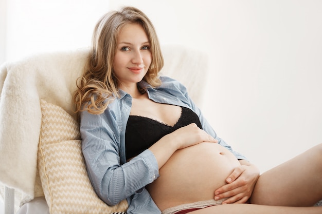 Close up portrait of young beautiful cheerful future mother with light curly long hair in comfortable home clothes smiling gently, lying on cozy bed, holding her pregnant belly with hands.