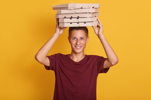 Free photo close up portrait of young cheerful delivery man with red t-shirt standing,holding stack of cardboard pizza boxes on grey wall.