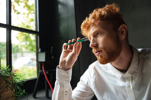 Free photo close-up portrait of young concentrated readhead bearded man, holding green pen, looking at window