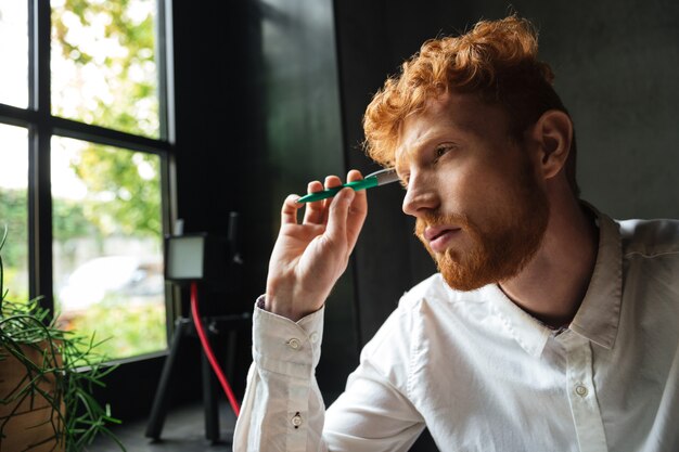 Close-up portrait of young concentrated readhead bearded man, holding green pen, looking at window