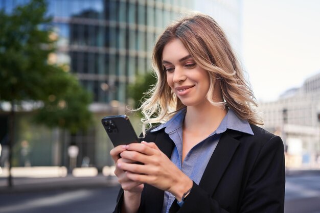 Close up portrait of young corporate woman in black suit holds smartphone texting message while stan