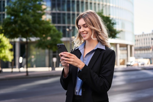 Free photo close up portrait of young corporate woman in black suit holds smartphone texting message while stan