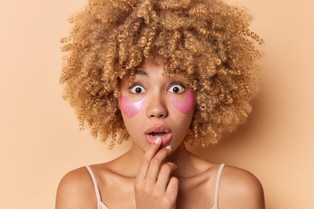 Close up portrait of young curly woman feels surprised keeps mouth opened applies pink collagen patches under eyes to reduce puffiness stands indoor against beige background. Cosmetic procedures