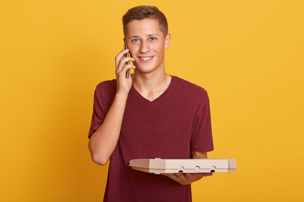 Close up portrait of young delivery man holding pizza boxes in hands and having conversation via mobile phone, wearing casually, looking smiling at camera, posing isolated on yellow.