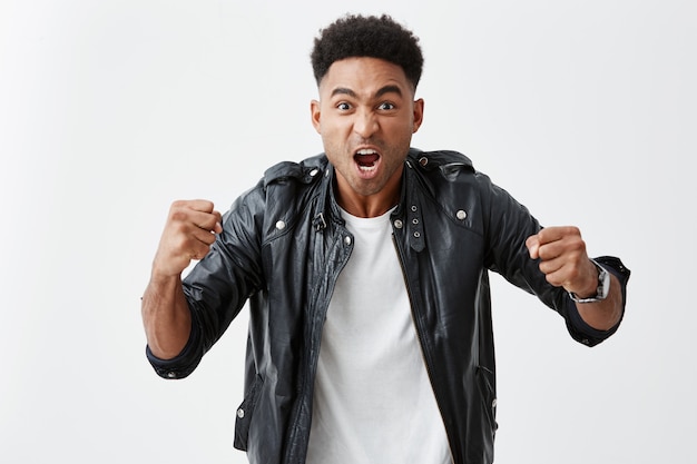 Close up portrait of young expressive dark-skinned young man with curly hair in white t shirt and leather jacket gesticulating with hands, screaming, cheering his favorite football team on stadium.