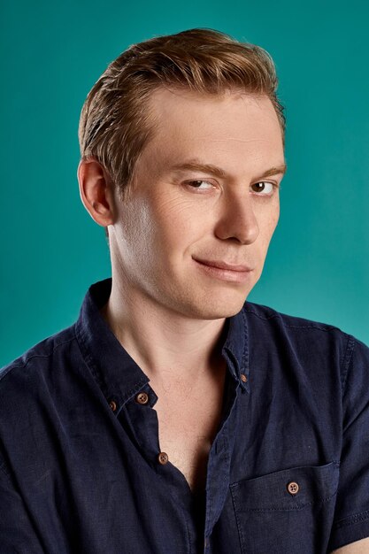 Close-up portrait of a young fashionable ginger male in a stylish navy t-shirt looking at the camera and smiling while posing on blue studio background. Human facial expressions. Sincere emotions conc