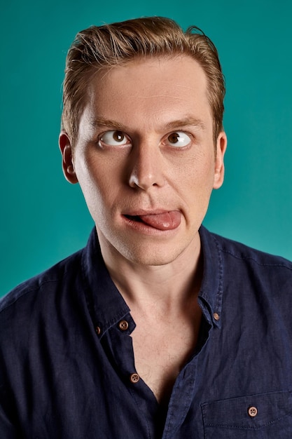 Close-up portrait of a young fashionable ginger man in a stylish navy t-shirt fooling around while posing on blue studio background. Human facial expressions. Sincere emotions concept. Copy space.