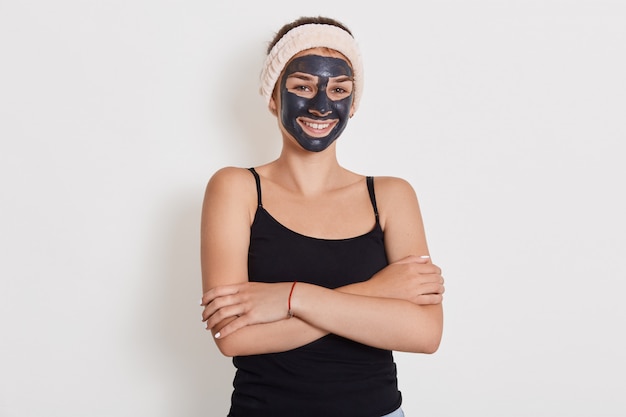 Close up portrait of young female applies homemade facial clay mask, has white hairband around head, smiles happily, keeps hands folded against white wall