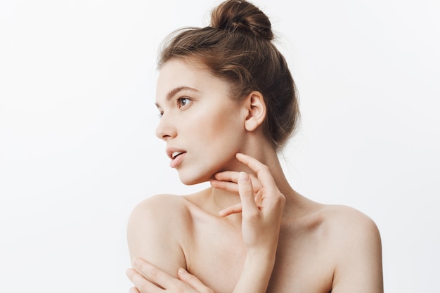 Free photo close up portrait of young female brunette student girl with bun hairstyle and baked shoulders looking aside with calm face expression touching jawline with fingers.