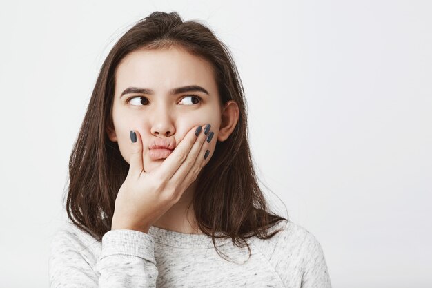 Close-up portrait of young female brunette with curious expression squeezing her face and thinking about something