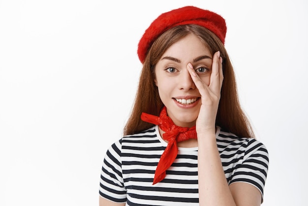 Ritratto ravvicinato di giovane studentessa in berretto francese che si tiene per mano sul viso toccando la pelle del viso pulita con trucco naturale sorridente in telecamera su sfondo bianco