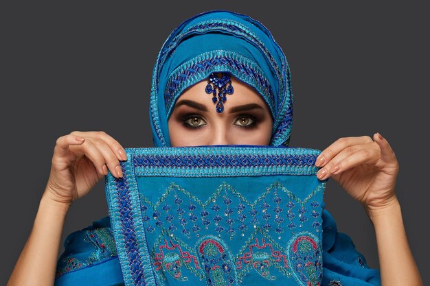Close-up portrait of a young female with smoky eyes wearing an elegant blue hijab decorated with sequins and jewelry. She is covering her face with a shawl and looking at the camera on a dark backgrou