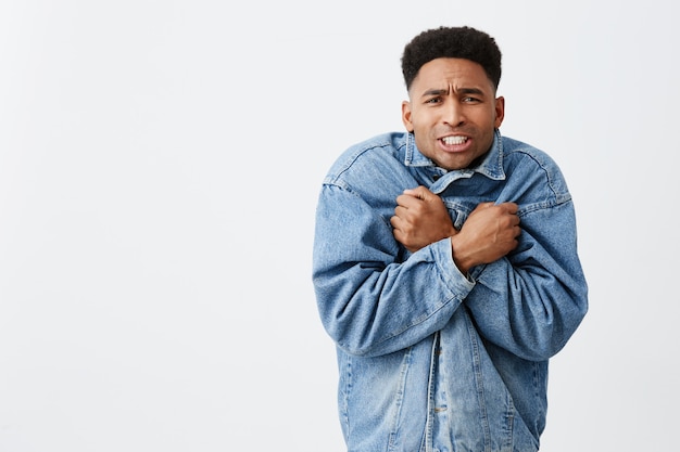 Close up portrait of young funny dark-skinned male with afro hairstyle in casual denim outerwear being frozen waiting friend outside in winter. Cold season.