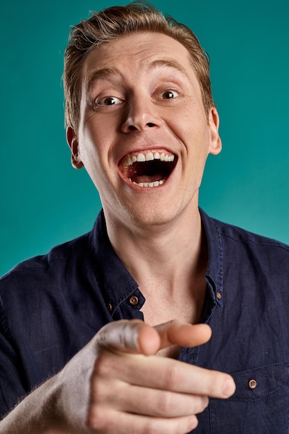 Free photo close-up portrait of a young funny ginger fellow in a stylish navy t-shirt looking at the camera and laughing at someone while posing on blue studio background. human facial expressions. sincere emoti