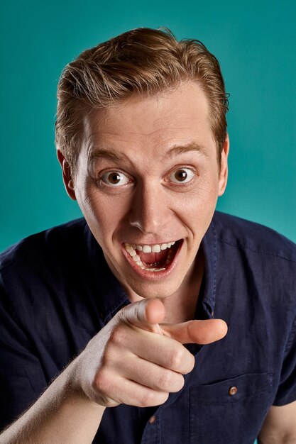 Close-up portrait of a young funny ginger guy in a stylish navy t-shirt looking at the camera and laughing at someone while posing on blue studio background. Human facial expressions. Sincere emotions