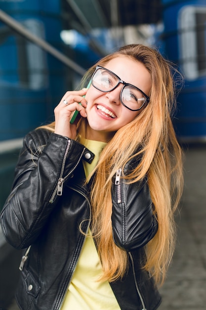 Close-up portrait of young girl with long hair in glasses outside in airport. She wears black jacket, speaking on phone and smiling