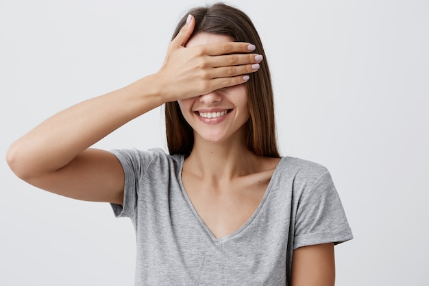 Close up portrait of young good-looking cheerful caucasian woman with dark long hair in gray t-shirt smiling with teeth, clothing eyes with hand, going to see her birthday present in few moments.