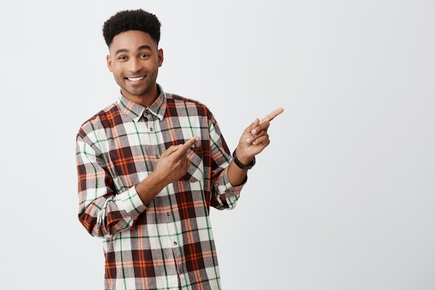 Close up portrait of young good-looking dark-skinned man with stylish dark afro hair in checkered shirt smiling with teeth, pointing aside won white wall with happy and joyful expression