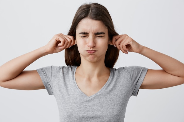 Chiuda sul ritratto di giovane bella ragazza caucasica castana divertente con capelli lunghi in camicia grigia casuale che tiene le orecchie con le mani, chiudendo gli occhi, facendo i fronti sciocchi per far ridere il piccolo bambino.