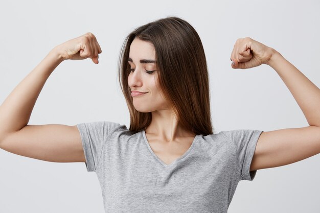 Close up portrait of young good-looking funny caucasian brunette girl with long hair in t-shirt playing with muscles, looking at them with confident and powerful face expression.