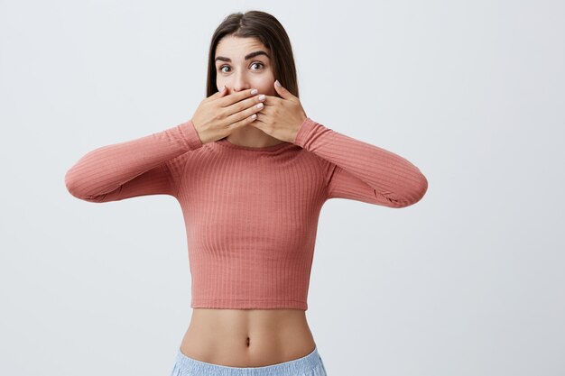 Close up portrait of young good-looking sexy caucasian female student with dark long hair in stylish outfit with naked belly looking  with happy expression, clothing mouth with both hands.