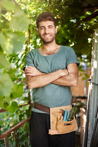 Ritratto ravvicinato di giovane giardiniere barbuto di bell'aspetto con attrezzi da giardino sorridente in piedi nell'ombra degli alberi che incrocia le mani guardando nella fotocamera con un'espressione facciale rilassata