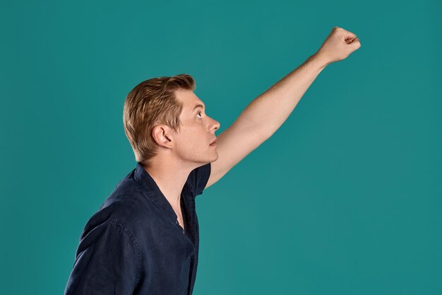 Close-up portrait of a young goodly ginger fellow in a stylish navy t-shirt acting like he is a superman while posing on blue studio background. Human facial expressions. Sincere emotions concept. Cop