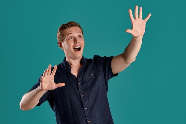 Close-up portrait of a young goodly ginger guy in a stylish navy t-shirt looking scared and acting like stopping someone while posing on blue studio background. Human facial expressions. Sincere emoti