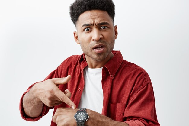 Close up portrait of young handsome black-skinned man with afro hairstyle in casual white t-shirt under red shirt pointing at hand watch with unsatisfied expression, after his friend being late.