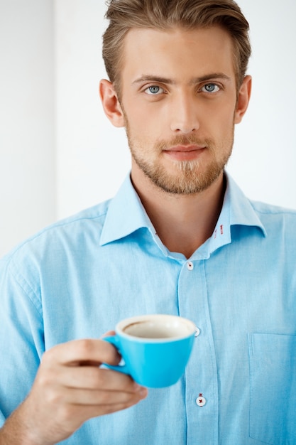 コーヒーカップを保持しているテーブルに立っている若いハンサムな自信を持って物思いにふけるビジネスマンの肖像画を閉じます。 。白い近代的なオフィスインテリア