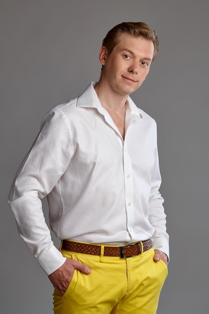 Close-up portrait of a young handsome ginger guy in a stylish white shirt and yellow trousers looking at the camera and smiling while posing on gray studio background. Human facial expressions. Sincer