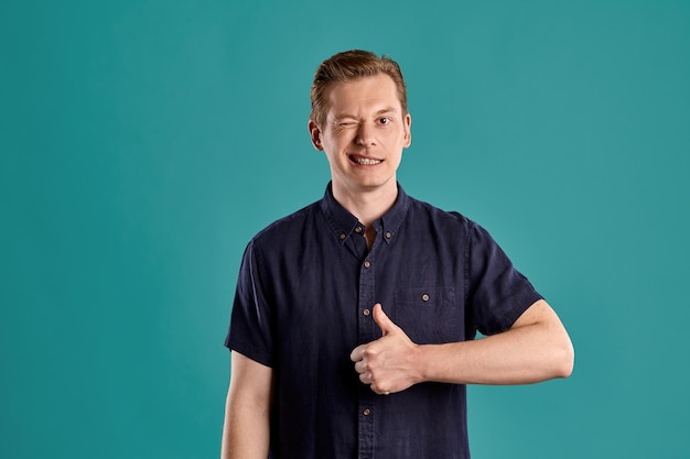 Close-up portrait of a young handsome ginger peson in a stylish navy t-shirt looking at the camera, winking and showing thumb up while posing on blue studio background. Human facial expressions. Since