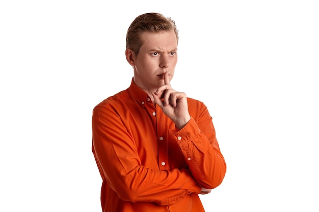 Крупным планом портрет молодого красивого рыжего человека в стильной оранжевой рубашке, смотрящего на кого-то и показывающего тихий знак, позируя изолированно на белом фоне студии. Выражение лица человека. С