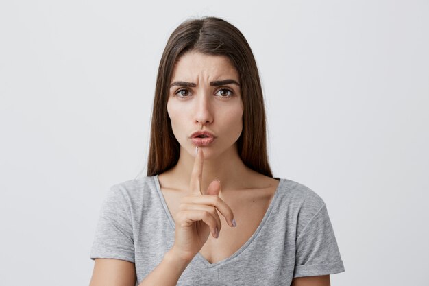 Close up portrait of young handsome sexy brunette caucasian woman with long hair in gray t-shirt holding index finger in front of lips, making hush gesture.