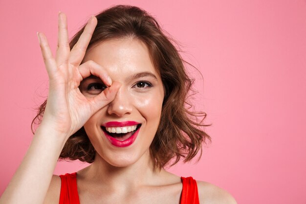 Close-up portrait of young happy girl with red lips  through OK sign