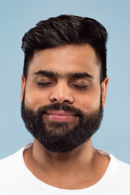 Close up portrait of young hindoo man with beard in white shirt isolated on blue background. Human emotions, facial expression, ad concept. Negative space. Dreaming with eyes closed.