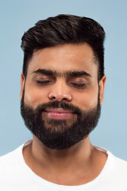 青い背景で隔離の白いシャツのひげを持つ若いヒンドゥー教の男の肖像画を閉じます。人間の感情、顔の表情、広告のコンセプト。ネガティブスペース。目を閉じて夢を見る。