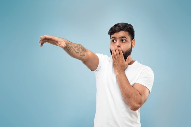 close up portrait of young indian man in white shirt. Pointing up being shocked and astonished.